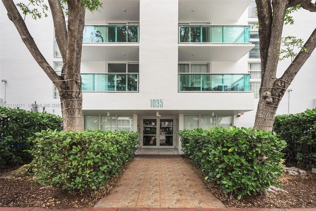 Exterior facade featuring white stucco finish, glass-railed balconies, a paved entryway, and mature landscaping