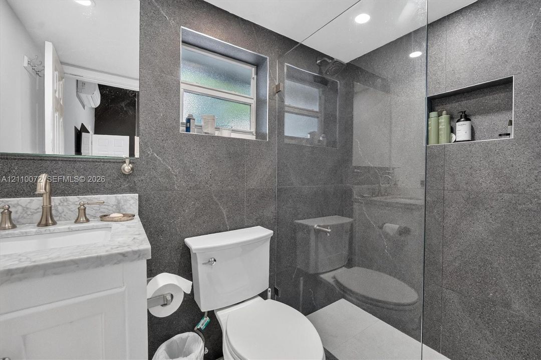 Bathroom featuring large-format dark stone wall tile, glass-enclosed shower with built-in recessed niche, stone-topped vanity with metallic faucet, and recessed lighting