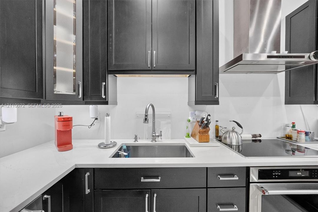 Dark finish cabinetry with glass-front accents, solid-surface countertops, stainless steel undermount sink, gooseneck faucet, and integrated stainless steel vent hood
