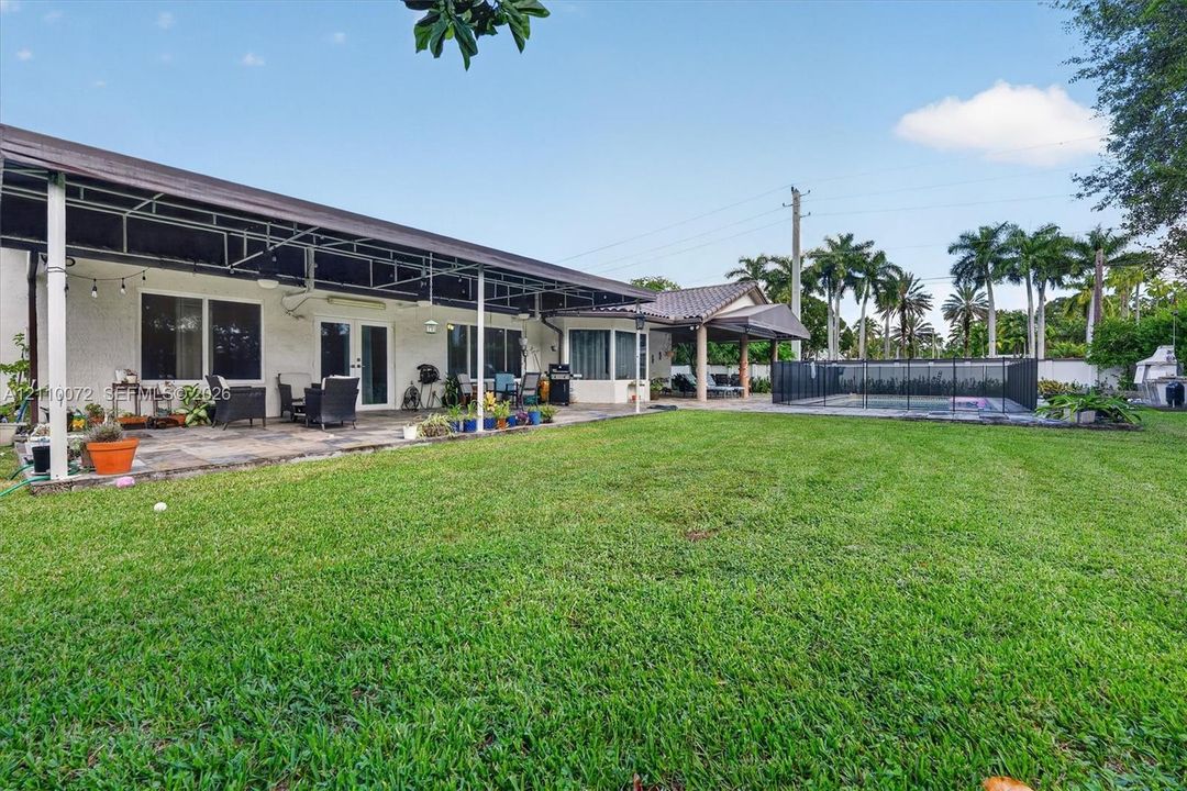 Backyard featuring an expansive lawn, stone-paved patio, covered terrace, in-ground swimming pool with safety fencing, and mature palm trees