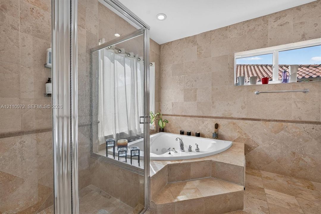 Bathroom featuring natural stone tile wall and floor surfaces, a raised corner jetted soaking tub, and a glass-enclosed shower with chrome fixtures