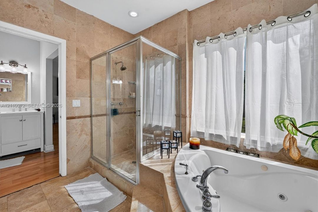 Full-sized jetted soaking tub with a tiled surround, glass-enclosed shower, natural stone wall and floor tile, recessed lighting, and a white vanity with an integrated sink