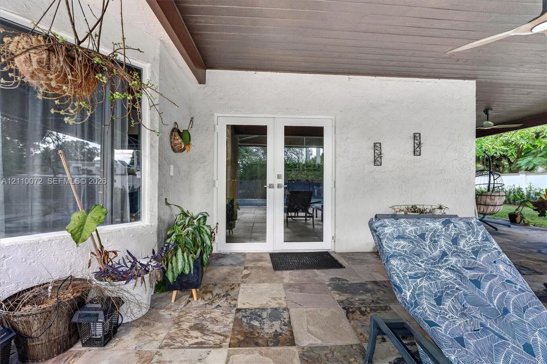 Covered patio featuring multi-tonal stone flooring, white textured exterior wall, dual-pane glass French doors, and a wood-finish ceiling with an integrated overhead fan
