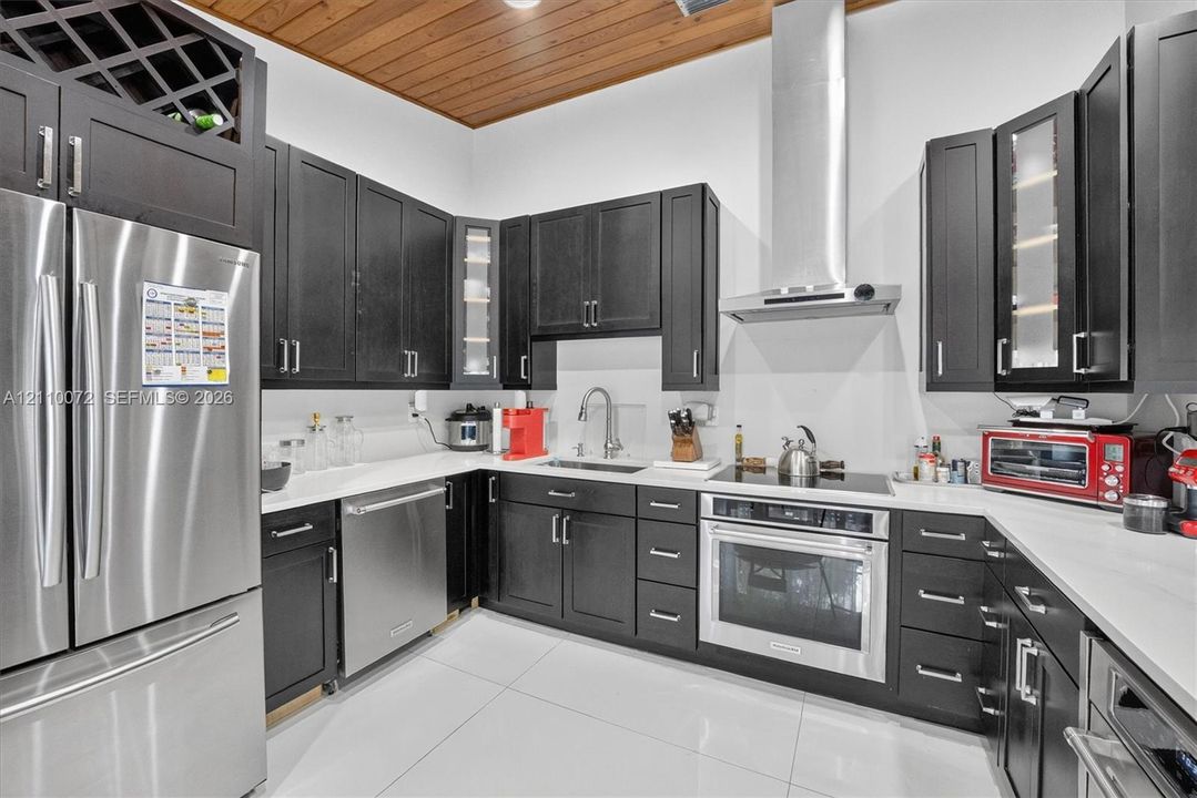 Kitchen featuring dark cabinetry, solid-surface countertops, wood-plank ceiling, stainless steel appliances, and a glass-front display cabinet