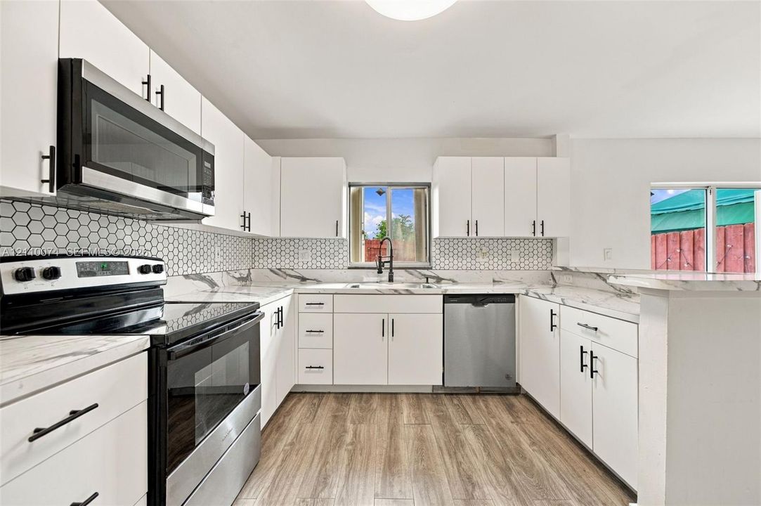 Kitchen featuring white cabinetry with matte black hardware, stone-patterned countertops, hexagonal tile backsplash, stainless steel appliances, and wood-finish flooring