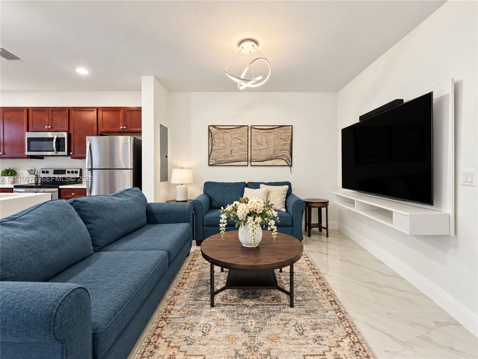 Open-concept space featuring large-format tile flooring, a wall-mounted floating media console, a modern geometric ceiling light fixture, and kitchen cabinetry paired with stainless steel appliances