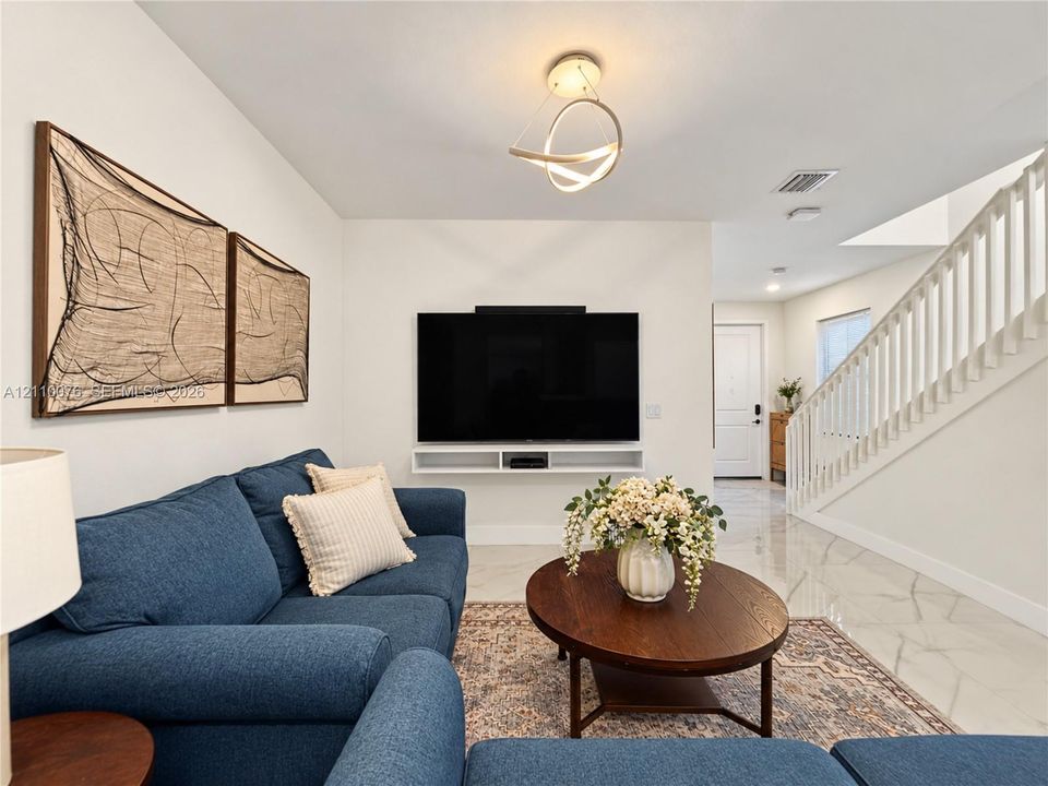 Open-concept interior featuring large-format tile flooring, a contemporary geometric ceiling light fixture, a wall-mounted floating shelf, and a white staircase with a wooden handrail