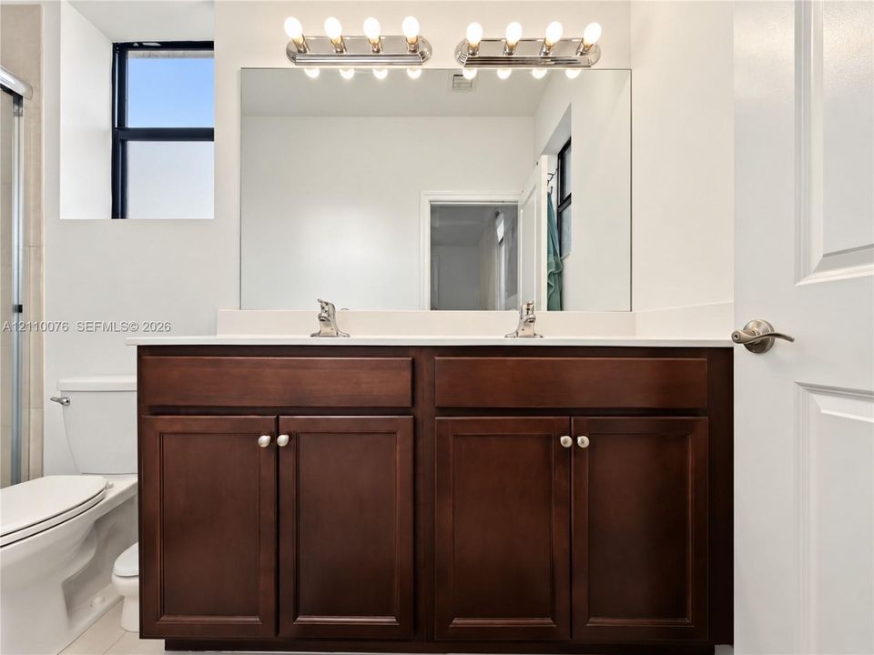 Double vanity featuring dark wood-finish cabinetry, a white countertop with integrated dual sinks, chrome fixtures, and a wall-to-wall mirror with multi-bulb overhead lighting