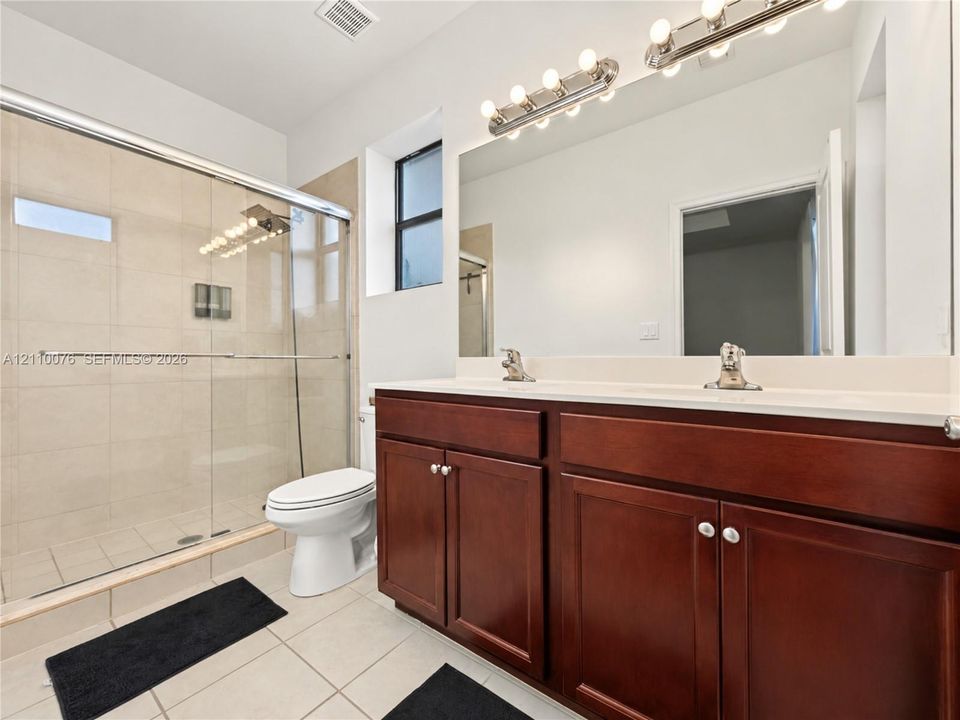 Dual-sink vanity with dark wood cabinetry and a solid-surface countertop, full-width wall mirror, and walk-in glass-enclosed shower with tile surround