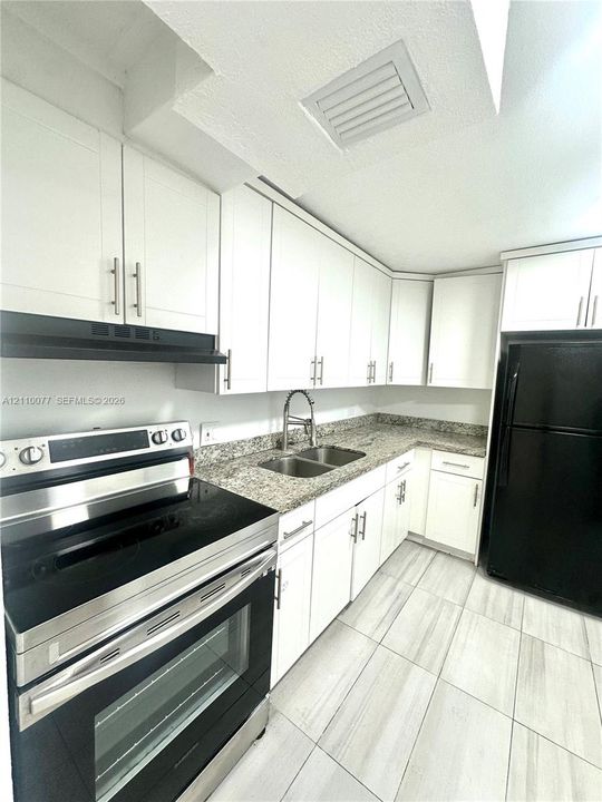 Kitchen featuring white shaker-style cabinetry, stone-finish countertops, dual-basin stainless steel sink with pull-down faucet, black kitchen appliances, and light-toned tile flooring