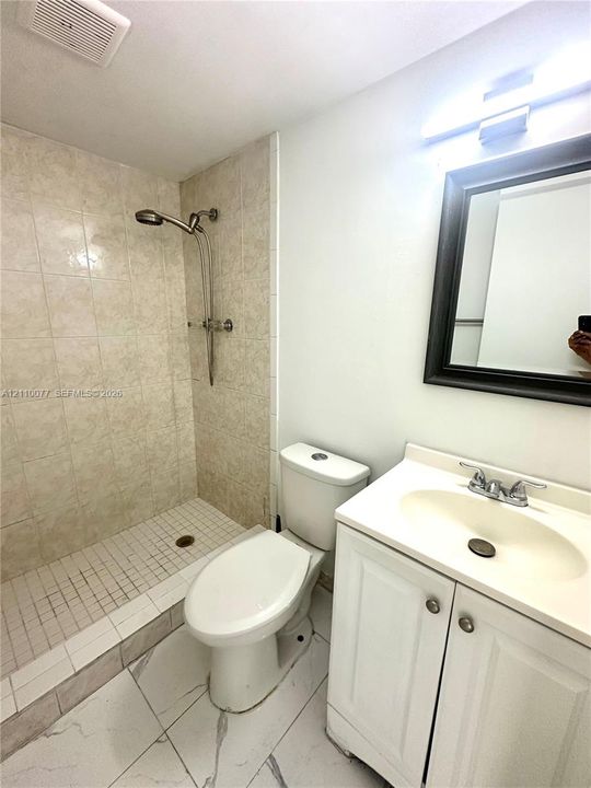 Bathroom featuring a walk-in shower with tiled surround, white vanity with integrated sink, over-mirror lighting, and large-format tile flooring with marble-style veining