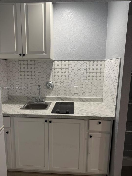 Kitchenette area featuring white shaker-style cabinetry, stone-patterned countertop, drop-in stainless steel sink with dual-handle faucet, integrated electric cooktop, and honeycomb-patterned tile backsplash