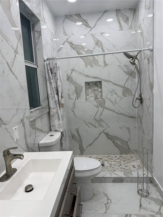 Full-height wall tiling with veined pattern, walk-in shower with pebble-tile flooring and recessed niche, floating vanity with integrated rectangular sink, and metallic finish faucet hardware