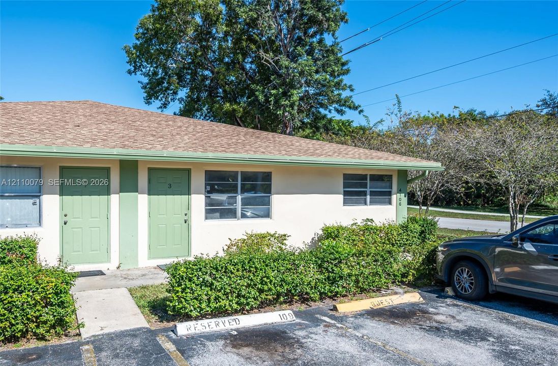 Single-story exterior featuring light-toned stucco, shingled roofing, dual entrance doors, and designated asphalt parking