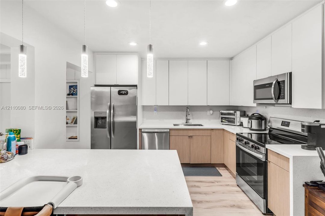 Kitchen featuring white upper cabinetry, light-toned wood-finish lower cabinetry, stone-composite countertops, stainless steel appliances, and minimalist pendant lighting