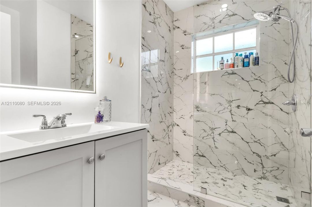 Bathroom featuring a stone-tile walk-in shower with a glass panel, vanity with an integrated sink, illuminated wall mirror, and polished chrome hardware