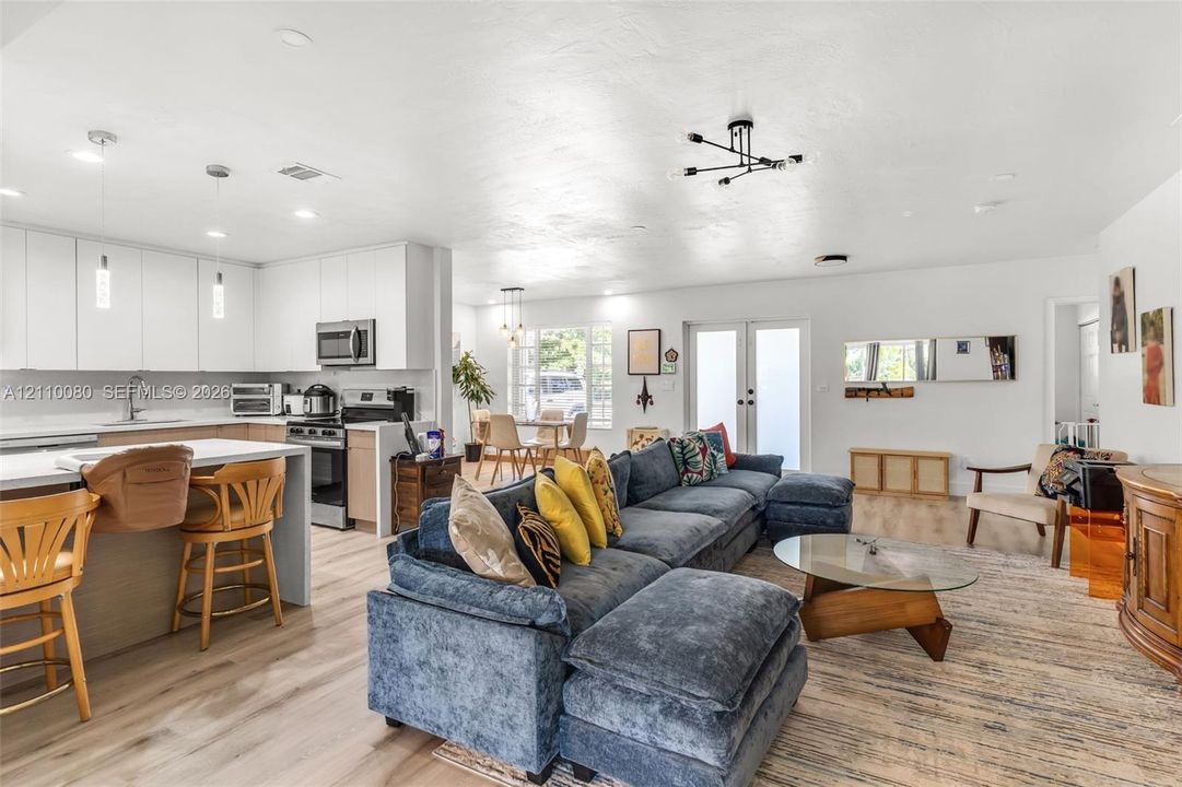 Open-concept living space featuring wood-finish flooring, white flat-panel cabinetry, a kitchen island with stone-finish countertops, modern multi-arm light fixtures, and glass-paneled double doors