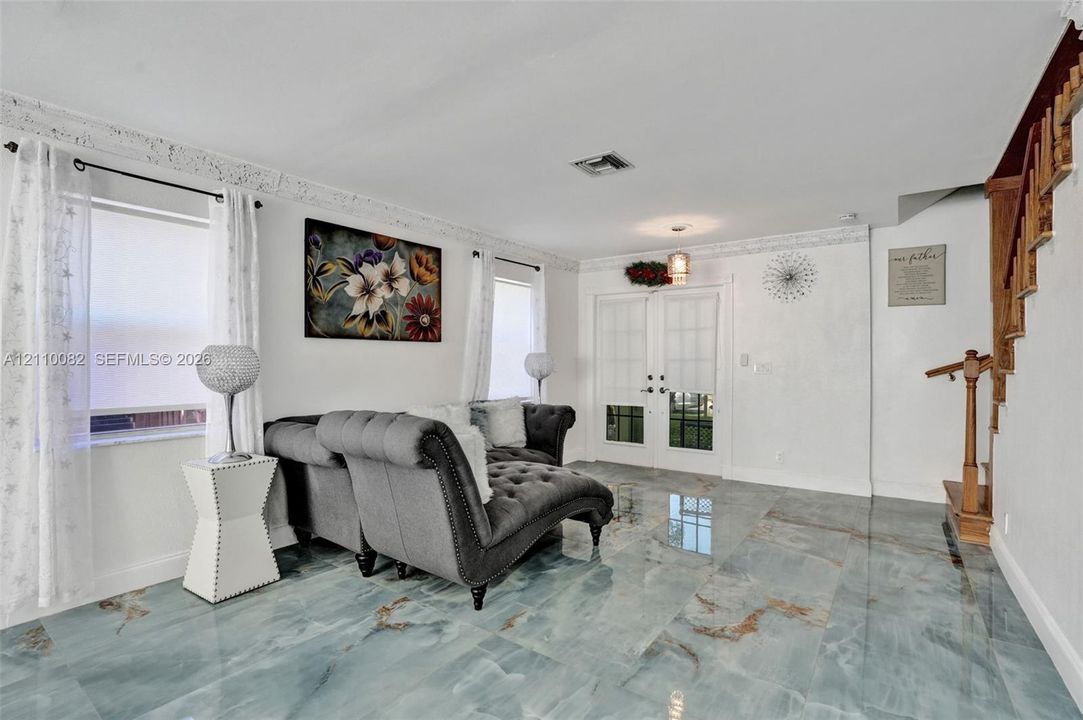 Polished stone flooring with natural veining, double-entry doors with glass paneling, decorative crown molding, and a wooden staircase featuring a stained handrail