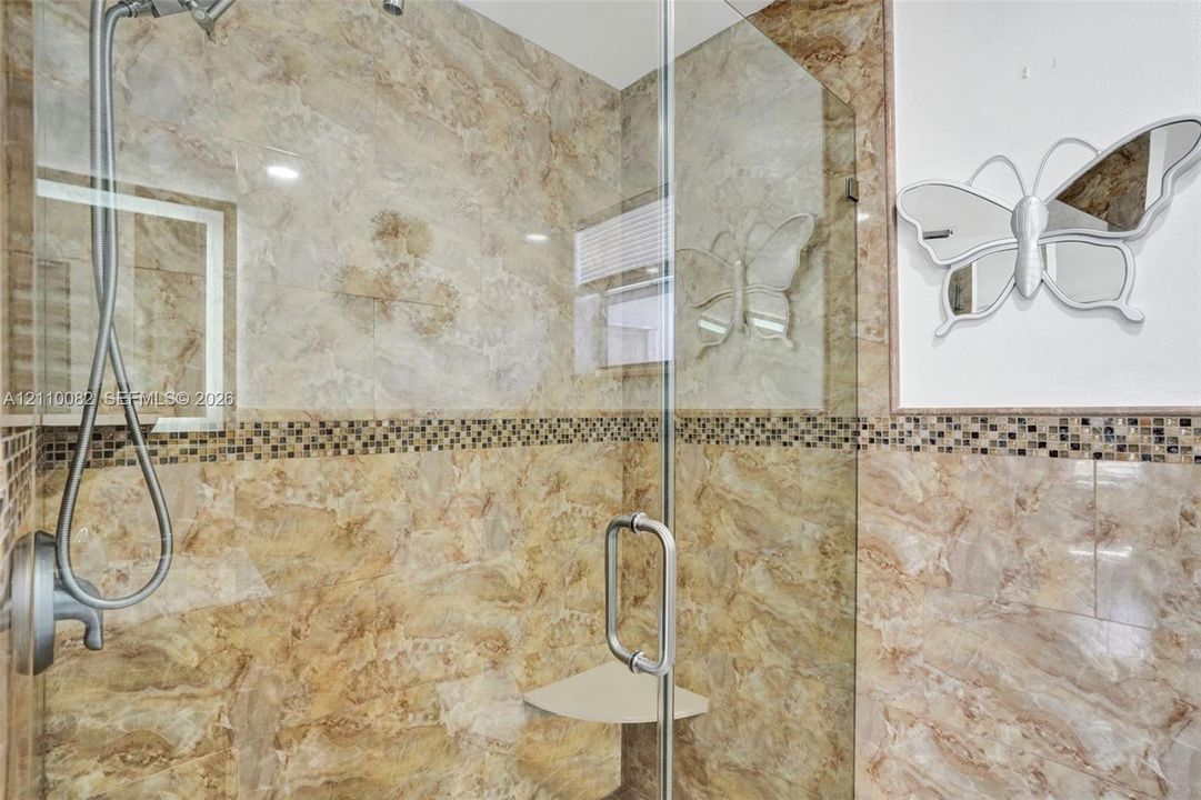 Walk-in shower featuring floor-to-ceiling stone-finish tile, glass enclosure, handheld shower fixture, built-in corner shelving, and decorative mosaic accent tile inlay