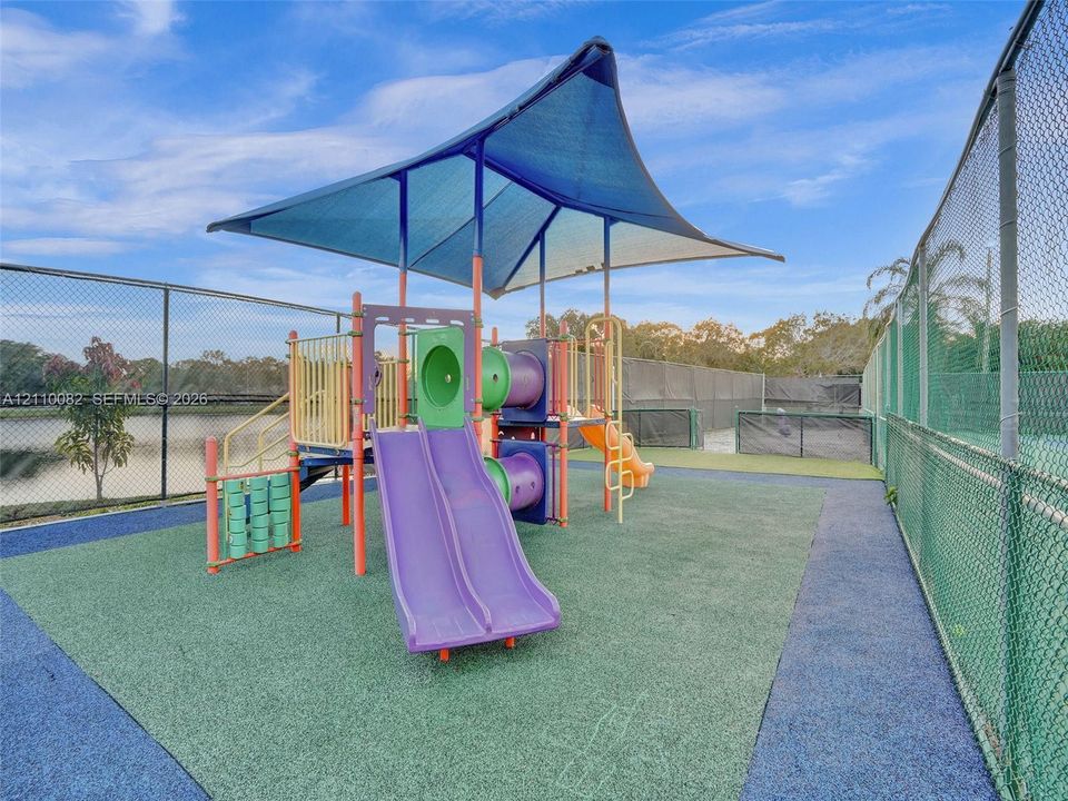 Recreational area featuring a playground structure with dual slides, a protective shade canopy, impact-absorbing surfacing, and perimeter chain-link fencing