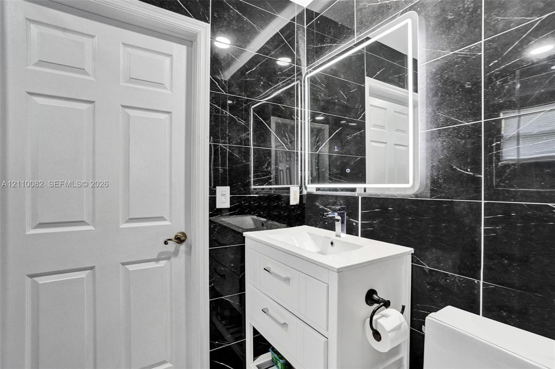 White vanity cabinet with an integrated basin, sleek chrome faucet, LED-backlit mirror, and full-height black stone-finish wall tile