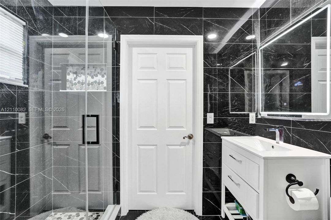 Bathroom featuring black, veined-stone wall tiling, glass-enclosed shower with a built-in recessed niche, white vanity with a rectangular sink, and an illuminated mirror