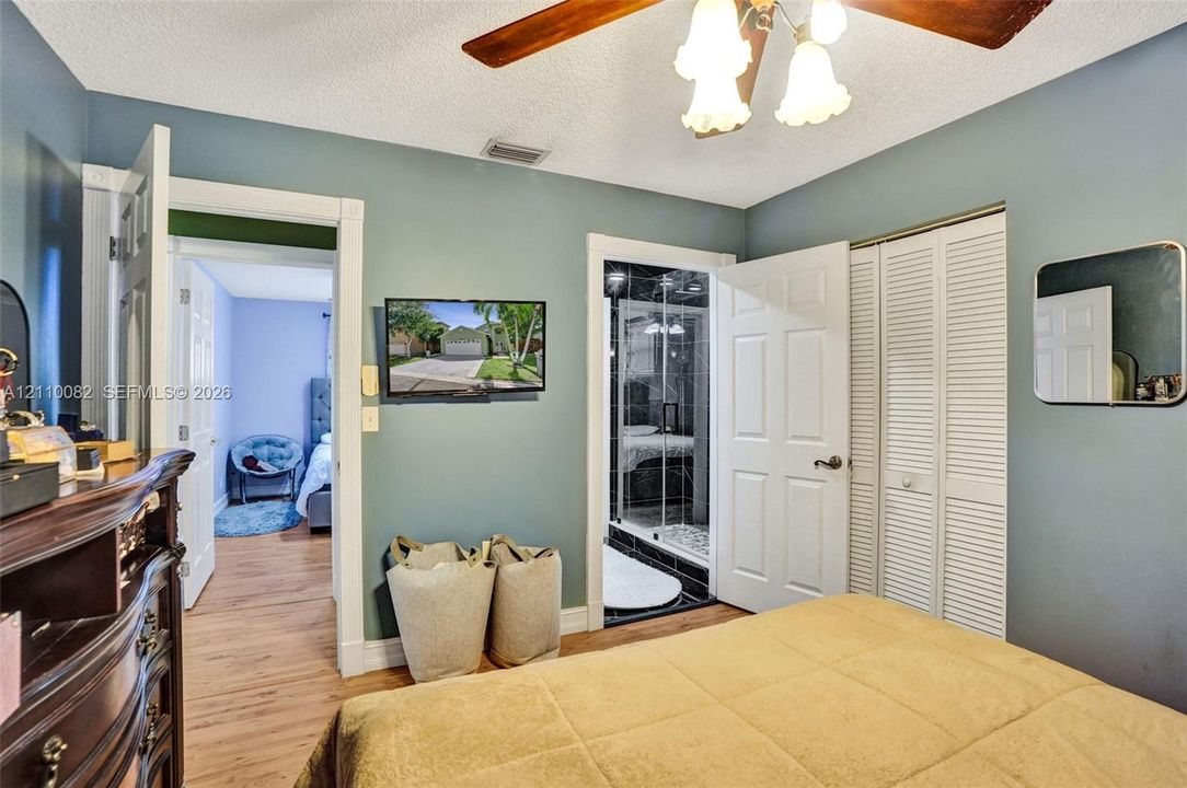 Room featuring wood-finish flooring, a multi-light ceiling fan, louvered bi-fold closet doors, and an attached bathroom with a glass-enclosed shower