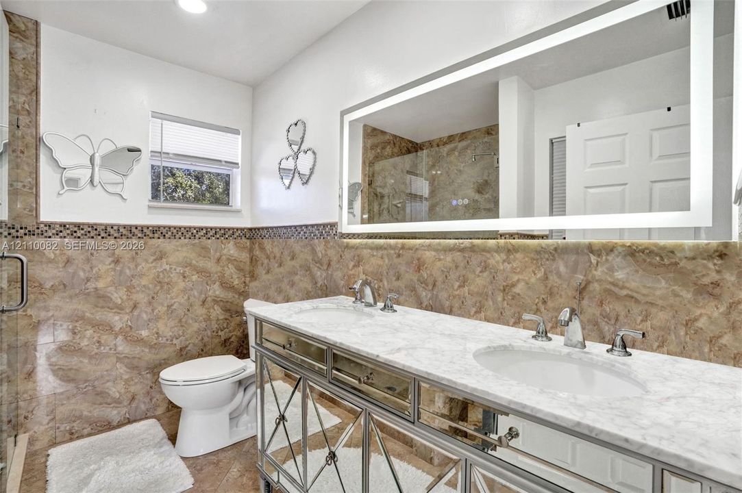 Double vanity with white stone countertop, dual undermount sinks, polished metal base, large LED-backlit mirror, and full-height stone tile wainscoting