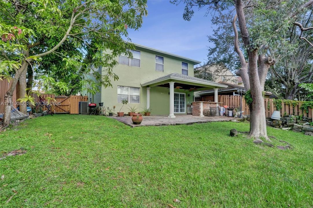 Rear exterior featuring a covered patio, built-in masonry outdoor kitchen, grass lawn, wood privacy fencing, and mature trees