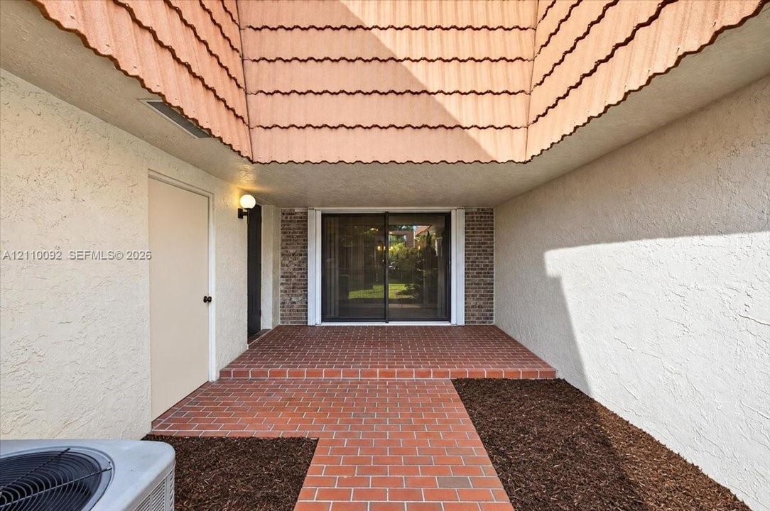 Covered entrance featuring a brick-paved walkway, stucco exterior walls, a recessed brick-accented doorway with sliding glass panels, and a decorative tile-shingle roof overhang