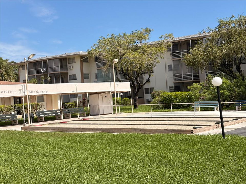 Outdoor shuffleboard court featuring a raked surface, covered seating area, perimeter lighting, and manicured lawn