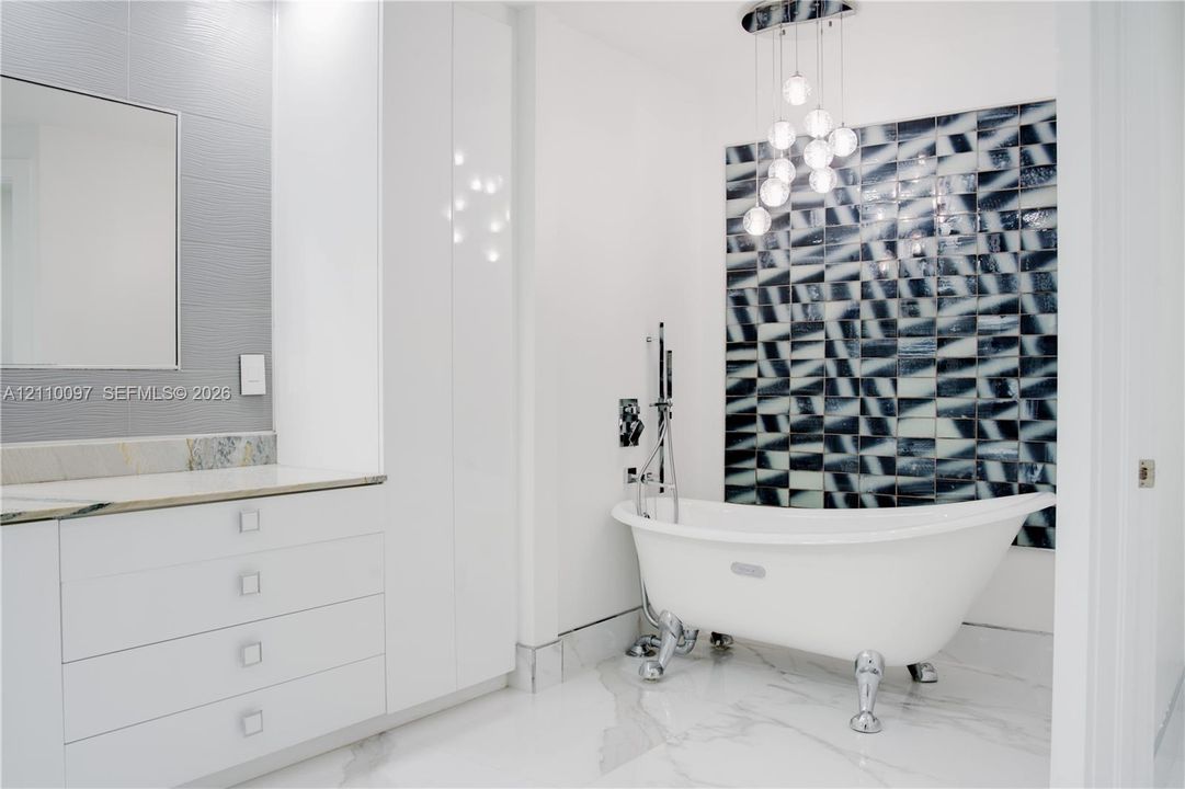 Clawfoot soaking tub with polished chrome hardware, geometric patterned accent wall, white cabinetry with integrated countertop, multi-globe pendant lighting, and large-format tile flooring