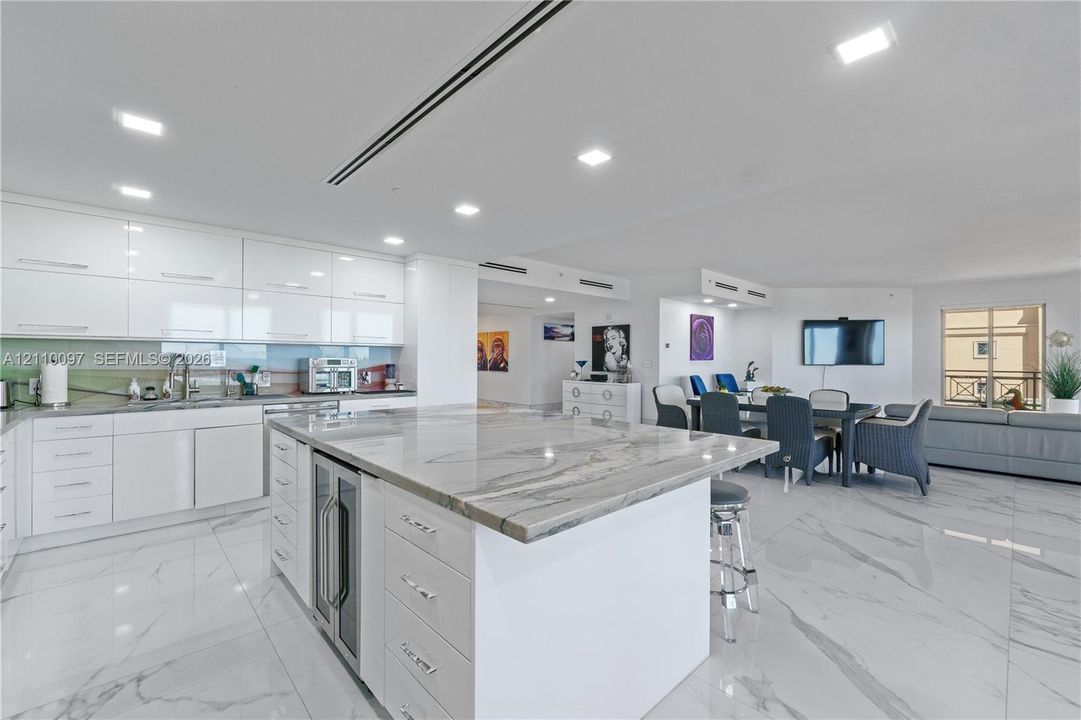 Kitchen featuring high-gloss white cabinetry, stone-finish waterfall island with built-in wine refrigeration, large-format tile flooring, and recessed ceiling lighting