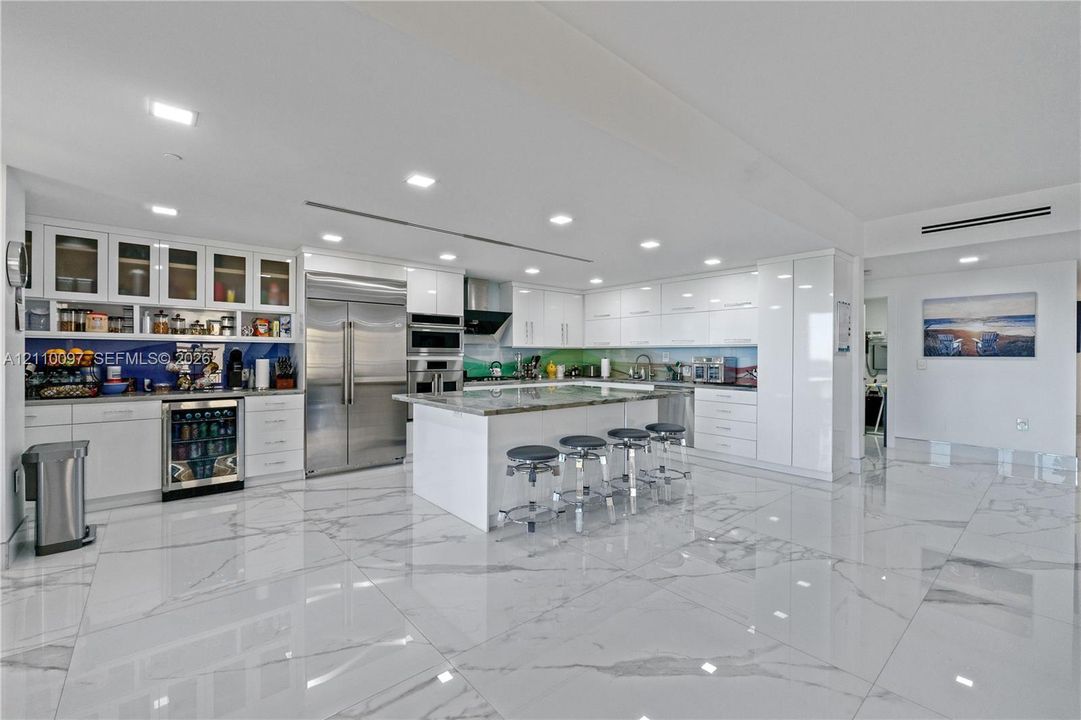 Modern kitchen featuring high-gloss white cabinetry, expansive stone-topped center island, large-format polished tile flooring, stainless steel appliances, and integrated wine refrigeration