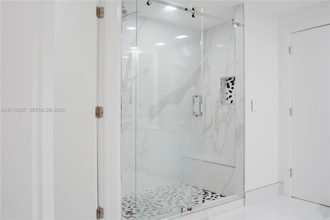 Walk-in shower featuring a frameless glass door, stone-finish wall tile with veining, pebble-style shower floor, built-in tiled bench, and an integrated recessed storage niche