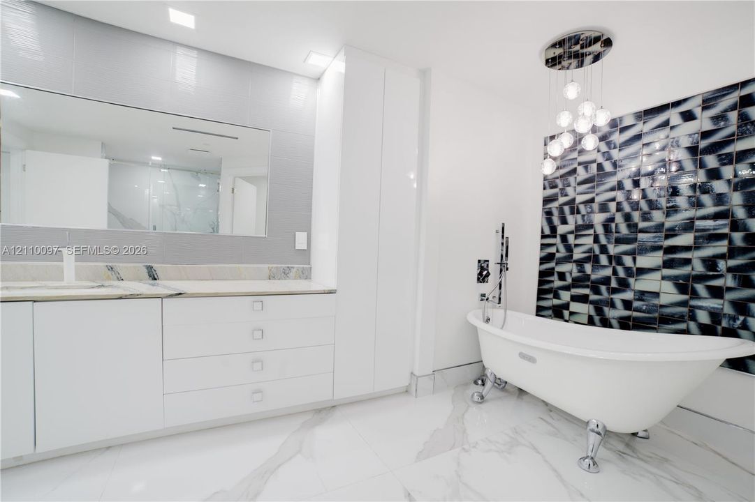 Freestanding clawfoot tub with chrome hardware, floor-to-ceiling geometric decorative wall tiling, custom white cabinetry with stone countertop, large-format veined flooring, and modern multi-globe chandelier lighting