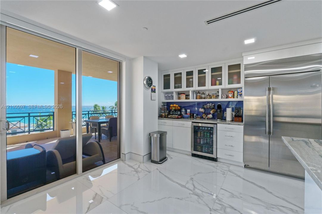 Large-format tile flooring, built-in white cabinetry with glass-front uppers, stainless steel side-by-side refrigerator, under-counter beverage cooler, and expansive floor-to-ceiling sliding glass doors opening to a covered balcony