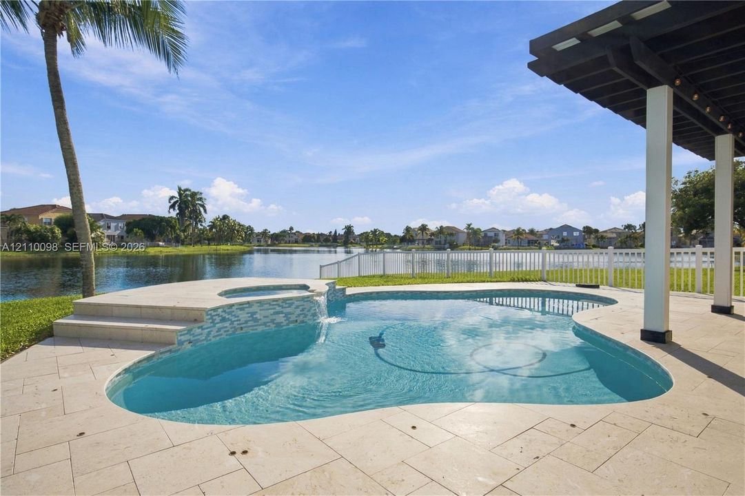 Pool area featuring a built-in spa with stone tile spillway, paved stone decking, covered patio structure, and perimeter fencing, set against a waterfront backdrop