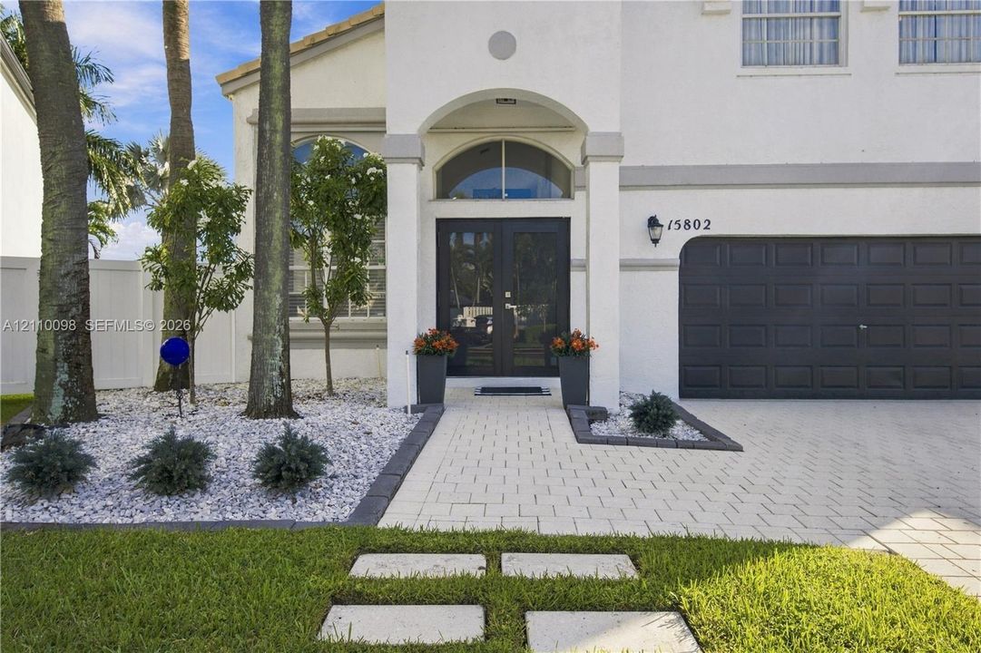 Stucco exterior featuring an arched entryway with double-door entry, a paver walkway, mature palm trees, and a multi-panel garage door