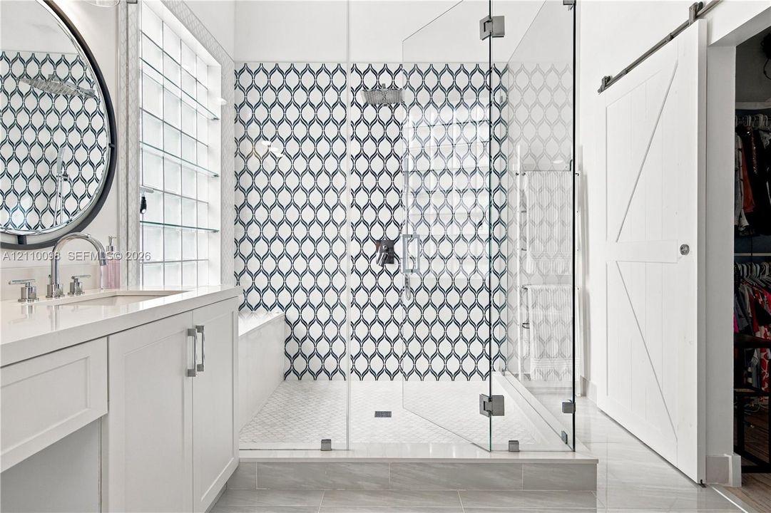 Walk-in shower featuring glass paneling and patterned tile surround, white cabinetry with light-toned countertop, round wall-mounted mirror, and white sliding barn door