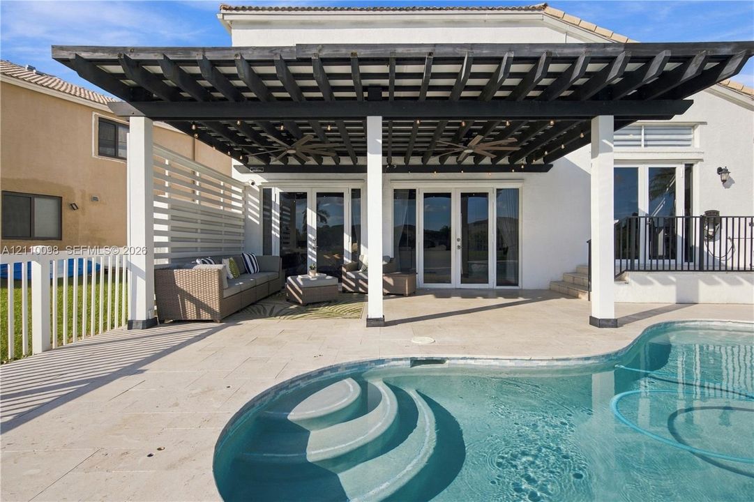 Covered patio featuring a dark-finish pergola, integrated ceiling fans, stone-finish decking, and a curved pool with recessed stairs