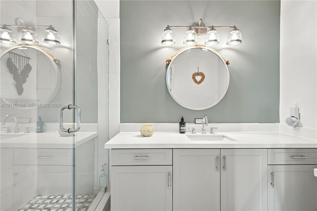 Bathroom featuring a vanity with light gray cabinetry, white solid-surface countertop, rectangular undermount sink, four-light vanity fixture, and a glass-enclosed shower with patterned tile flooring