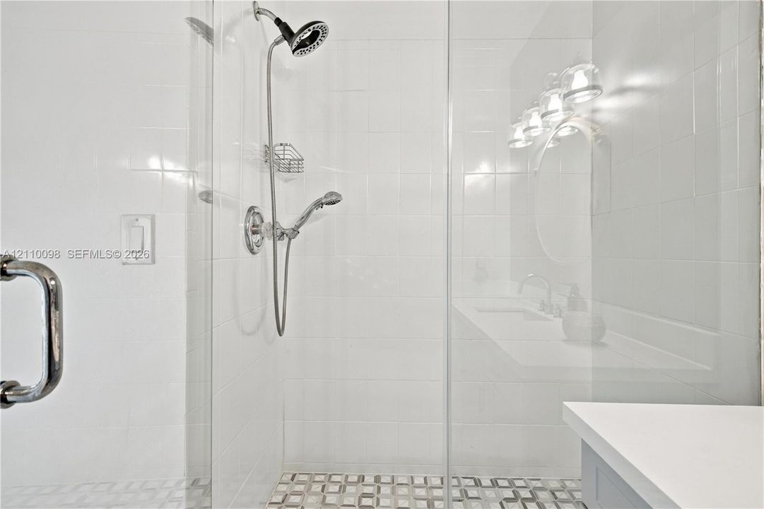 Walk-in glass shower enclosure featuring white tile walls, a handheld showerhead, built-in wire shelving, and a patterned tile floor