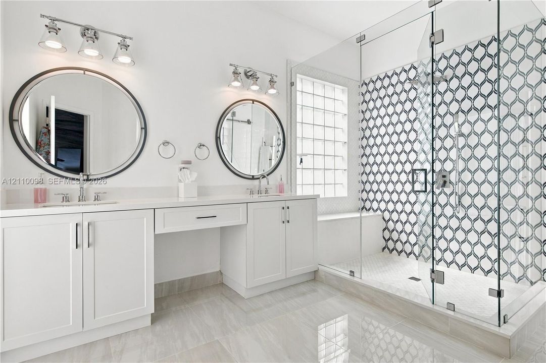 Double vanity featuring white cabinetry, oval mirrors, dual three-light fixtures, and a walk-in glass-enclosed shower with patterned tile surround