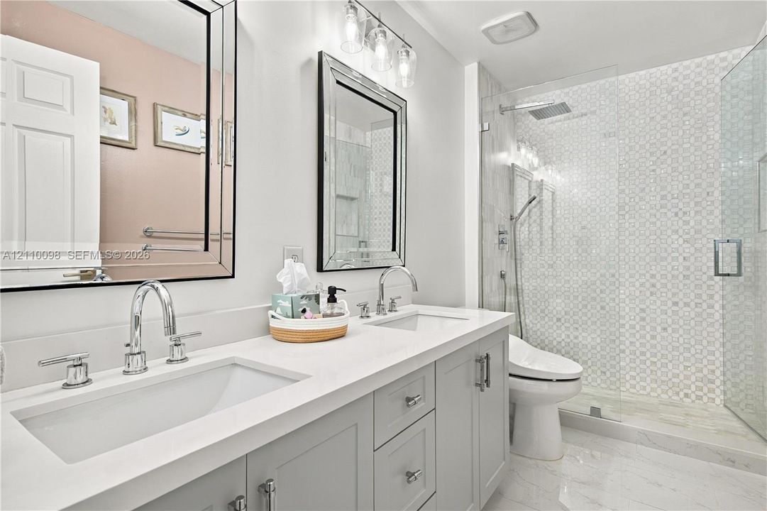 Dual-vanity cabinetry with a light-toned solid surface countertop, undermount sinks, polished chrome hardware, walk-in glass shower with decorative wall tile, and framed wall mirrors
