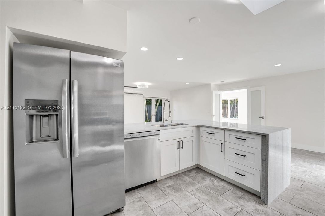 Kitchen featuring white shaker-style cabinetry, stainless steel appliances, quartz countertops, a waterfall-edge peninsula, and stone-tile flooring