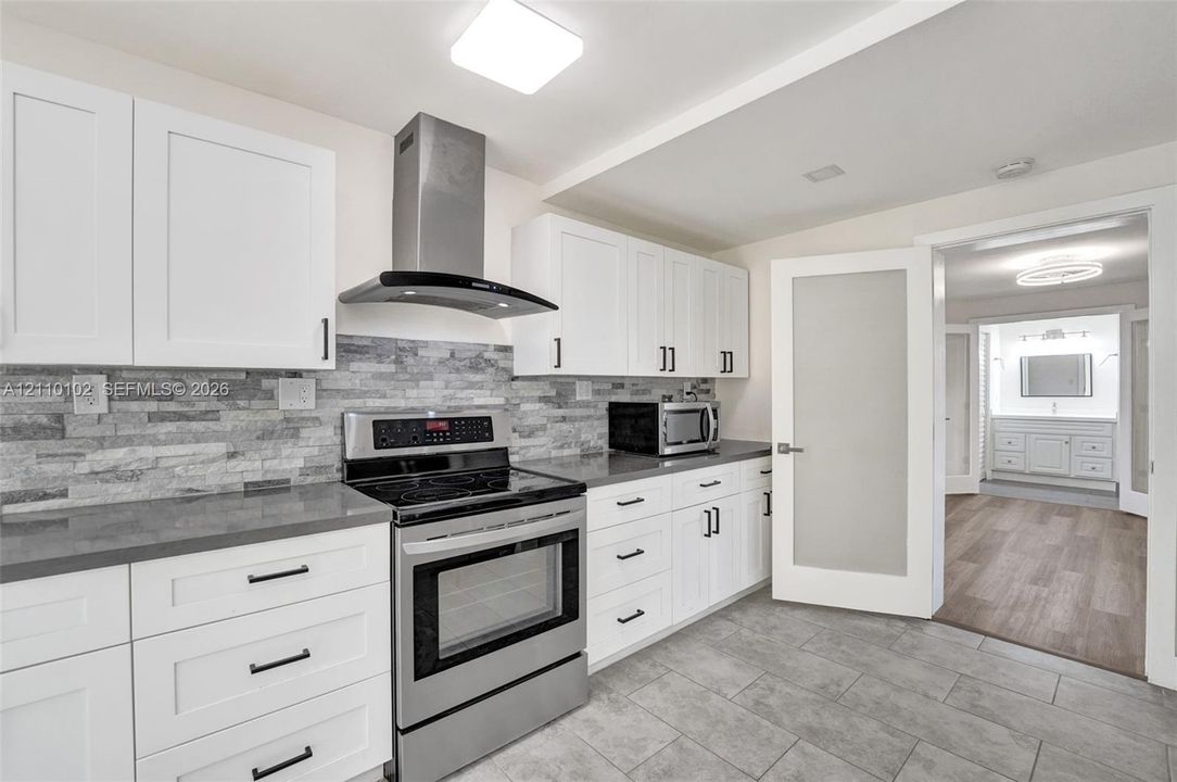 Kitchen featuring white shaker cabinetry, stainless steel appliances including a range and range hood, stone-finish backsplash, solid-surface countertops, and tiled flooring
