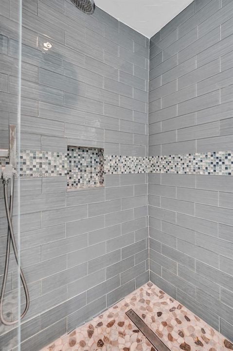 Walk-in shower featuring grey subway-style wall tile, a decorative glass mosaic accent band, a built-in recessed wall niche, a pebble-stone floor, and a handheld shower fixture