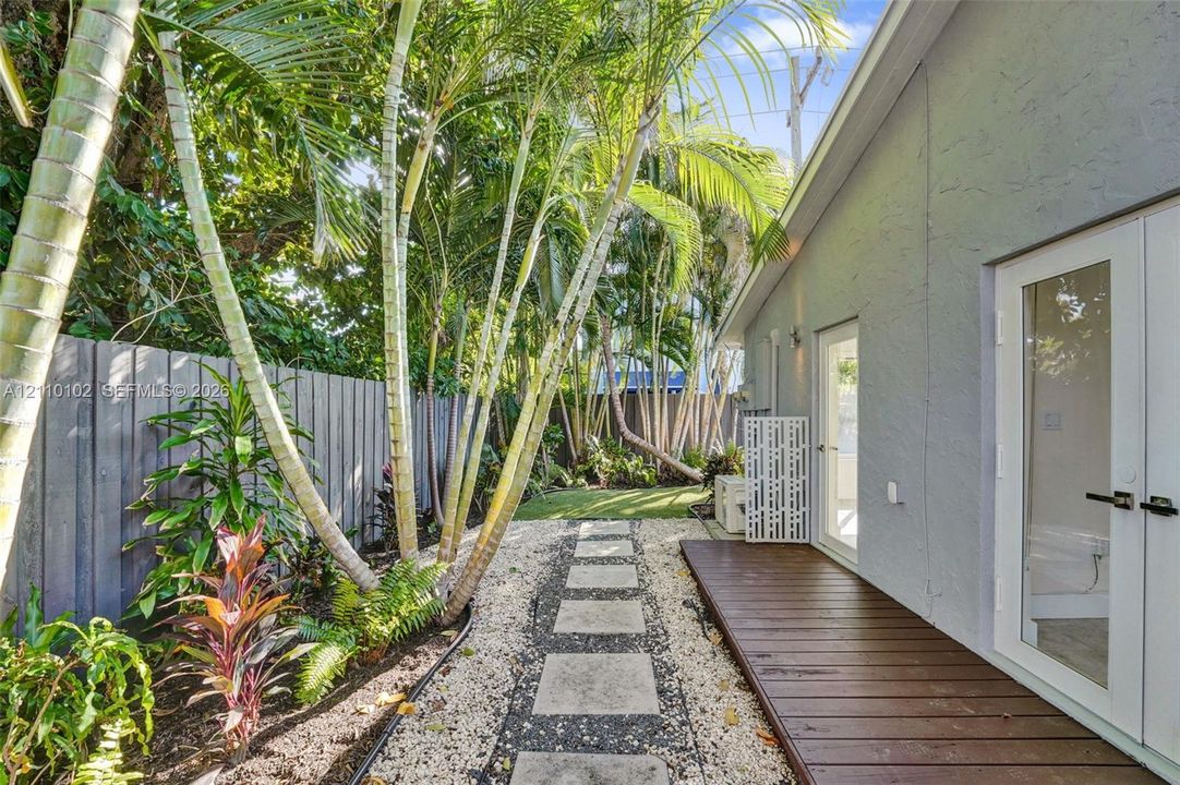 Exterior side yard featuring a wood-finish deck, a stone paver walkway, decorative gravel, vertical wood fencing, and mature palm trees