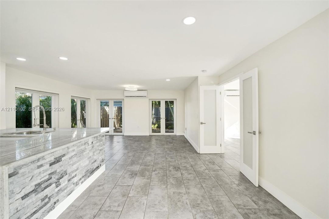Open-concept interior featuring large-format tile flooring, stone-veneer kitchen island base, recessed lighting, wall-mounted climate control unit, and glass-paneled entry doors